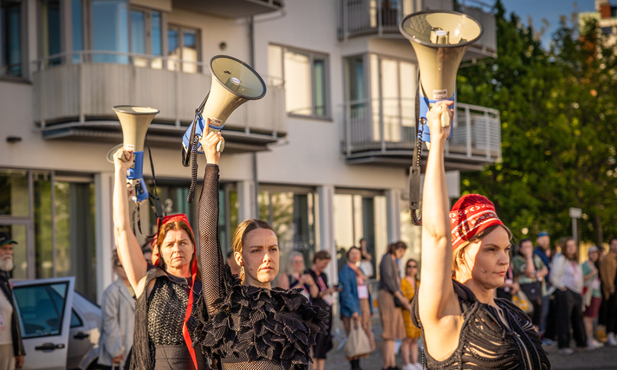 Three women in black costume, red hats and megaphones in the air outside surrounded by audience.