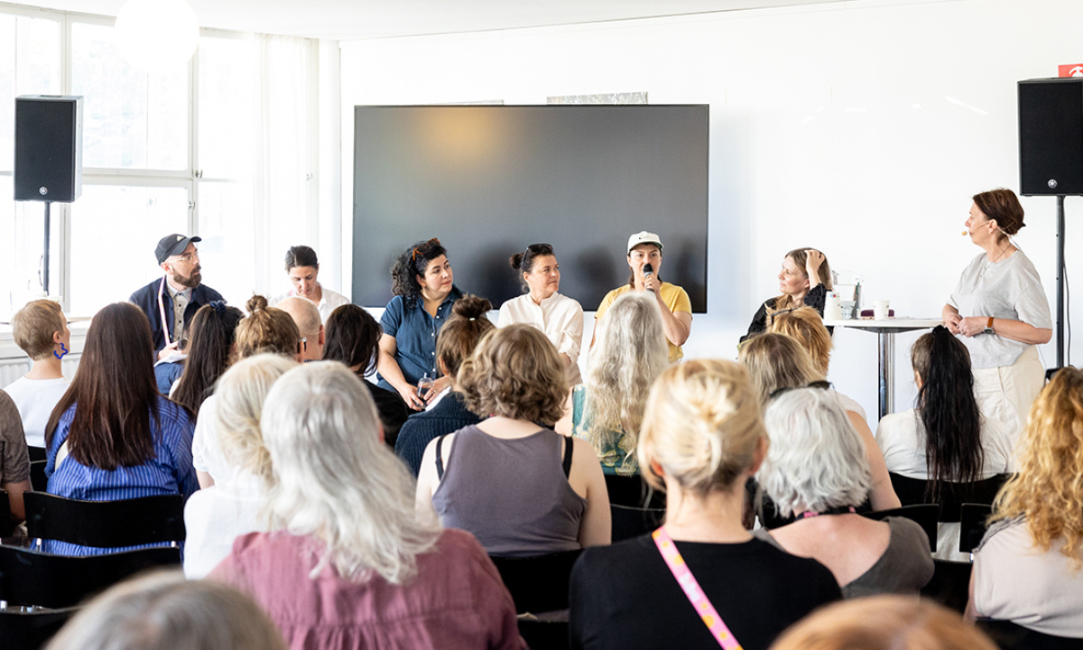 Flera personer sitter bredvid varandra på en scen för att ge ett seminarier. Vi ser publiken bakifrån.