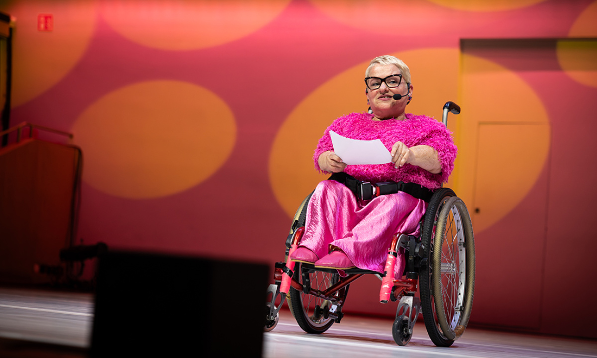 Kvinna kädd i rosa på en scen. Hon använder rullstol.